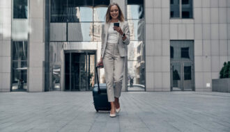 Office Style Diary - Businesswoman in light suit walking with luggage and smartphone outside modern office building, smiling while checking mobile device