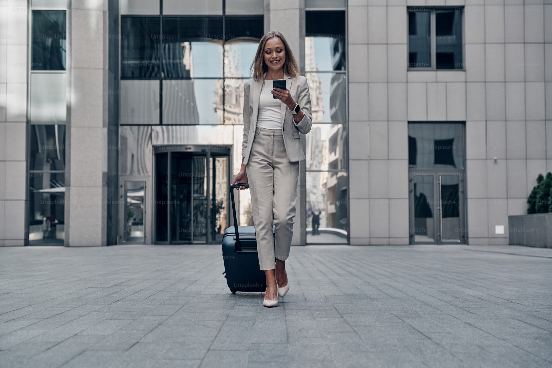 Office Style Diary - Businesswoman in light suit walking with luggage and smartphone outside modern office building, smiling while checking mobile device