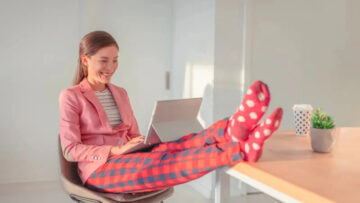 Office Style Diary - A cheerful woman in a stylish office setting, wearing a pink blazer and patterned red pants, with her feet up on the desk in fun, polka-dotted socks, working on a laptop.