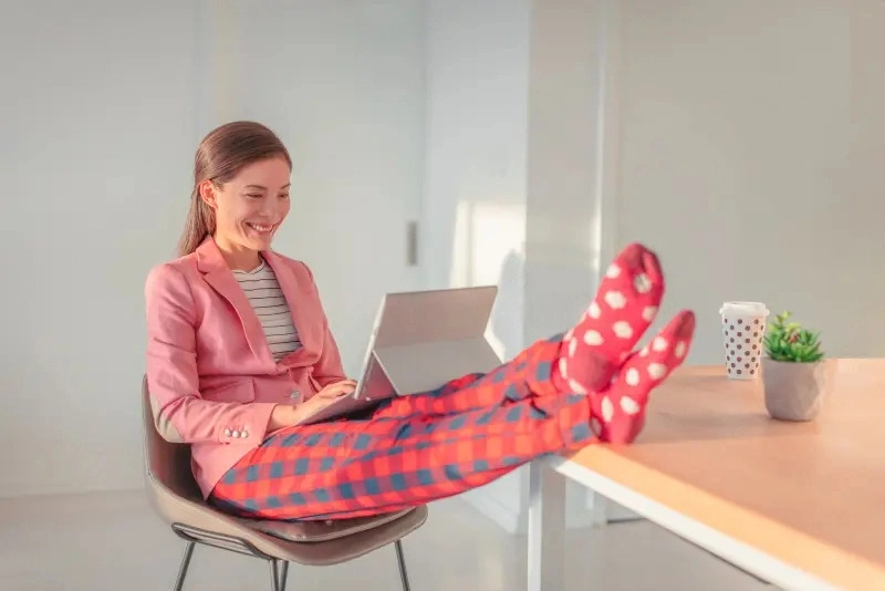 Office Style Diary - A cheerful woman in a stylish office setting, wearing a pink blazer and patterned red pants, with her feet up on the desk in fun, polka-dotted socks, working on a laptop.